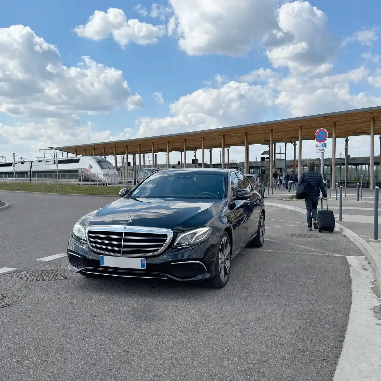 Une Mercedes Classe E noire à la dépose de la gare Lorraine TGV, sous sa longue toiture de bois ; un voyageur de dos part vers les quais avec sa valise