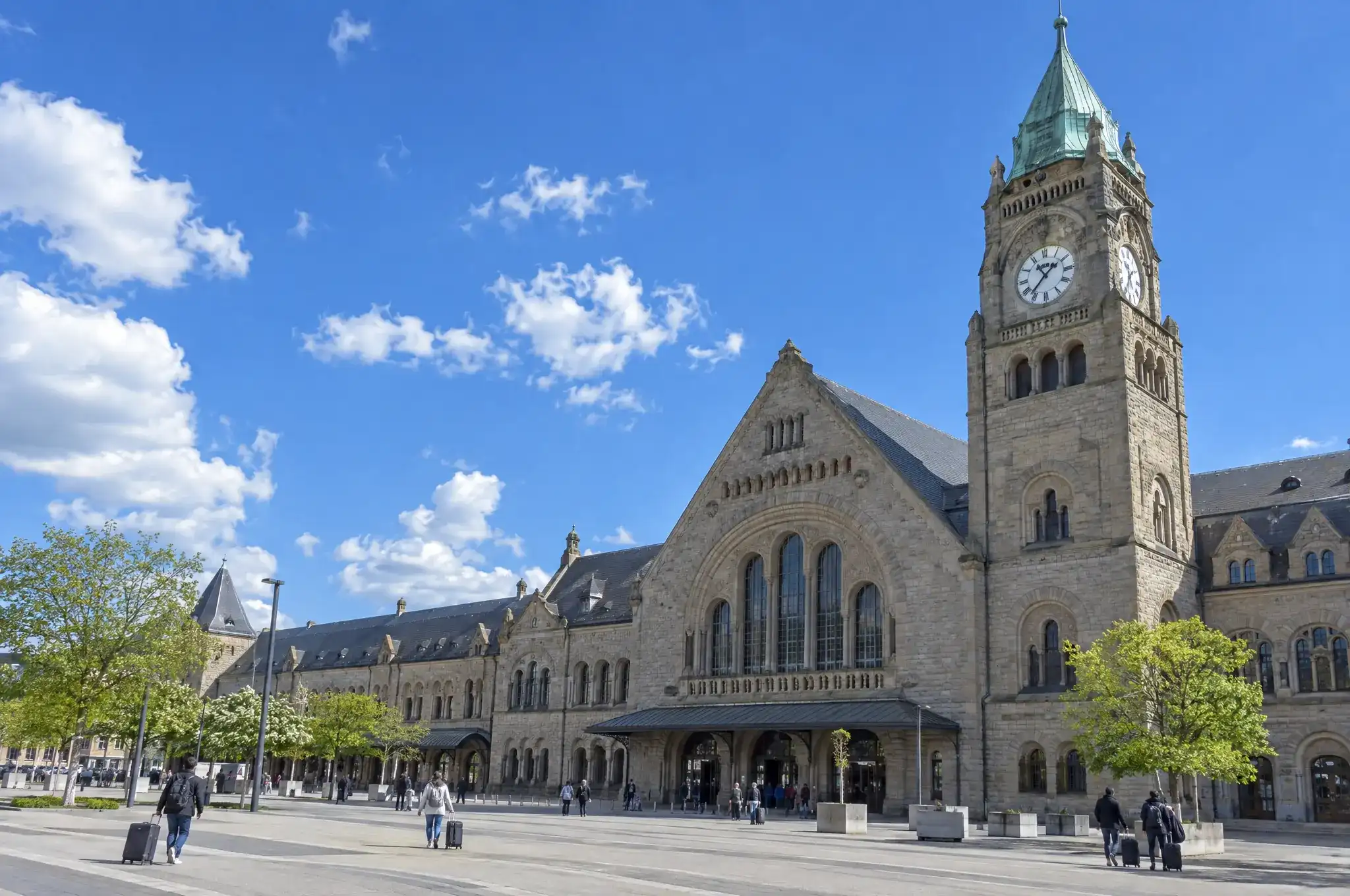 Le campanile à horloge d’une grande gare de pierre, voyageurs et valises sur le parvis, ciel bleu