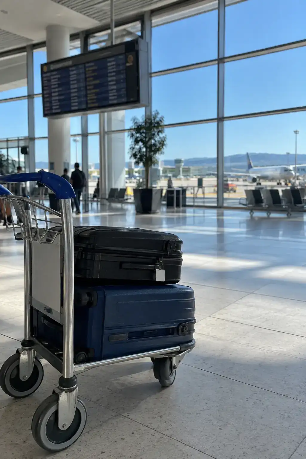 Hall des départs d’un aéroport, un chariot chargé de deux valises devant de grandes baies vitrées