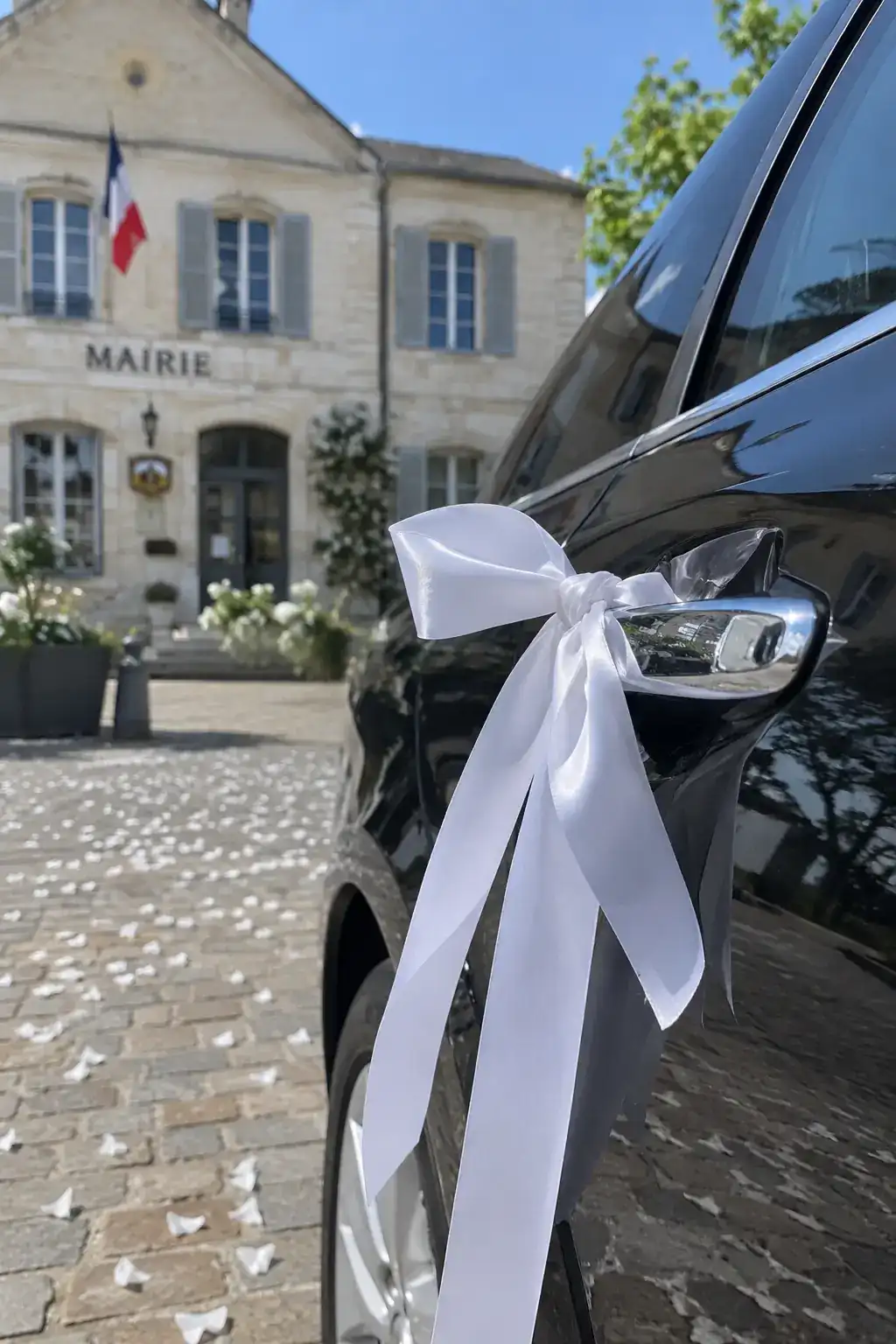 Un ruban de satin blanc noué sur la poignée d’une portière noire, devant une mairie de village en pierre claire