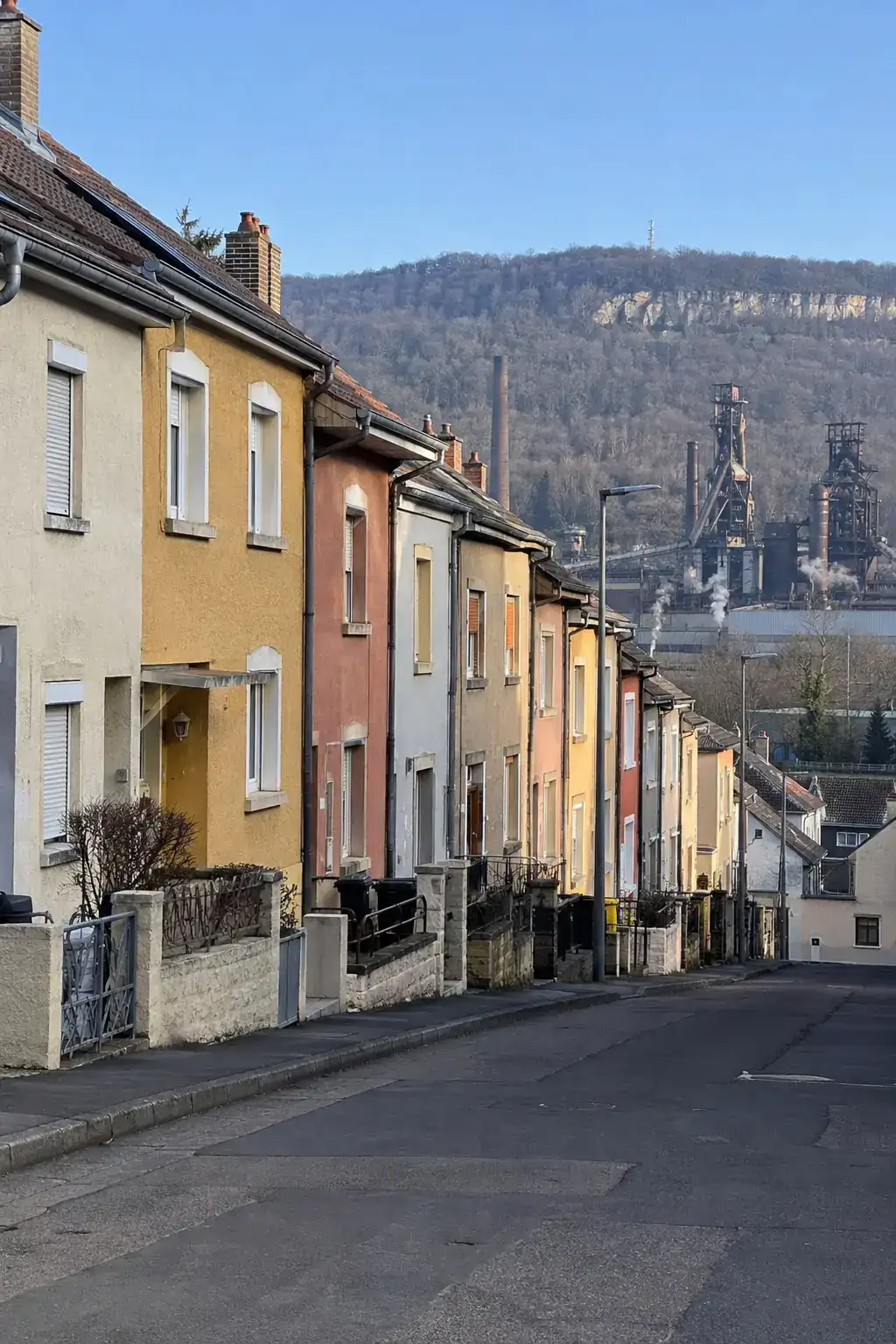 Une rue de maisons ouvrières mitoyennes aux enduits colorés, collines boisées et anciennes installations sidérurgiques au loin, ciel d’hiver