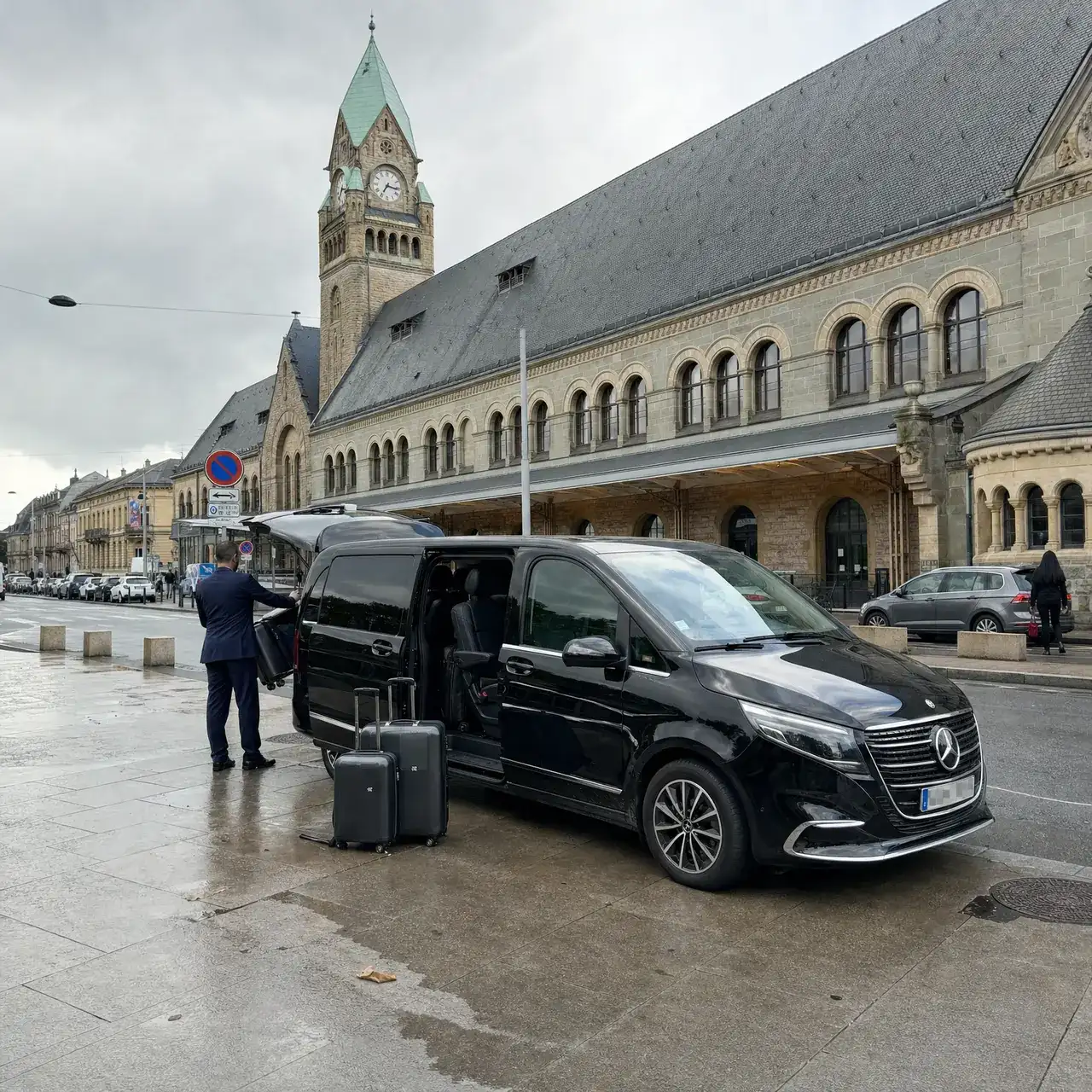 Une Mercedes Classe V noire, portière coulissante ouverte, devant la gare de Metz et son campanile ; un chauffeur de dos charge les valises