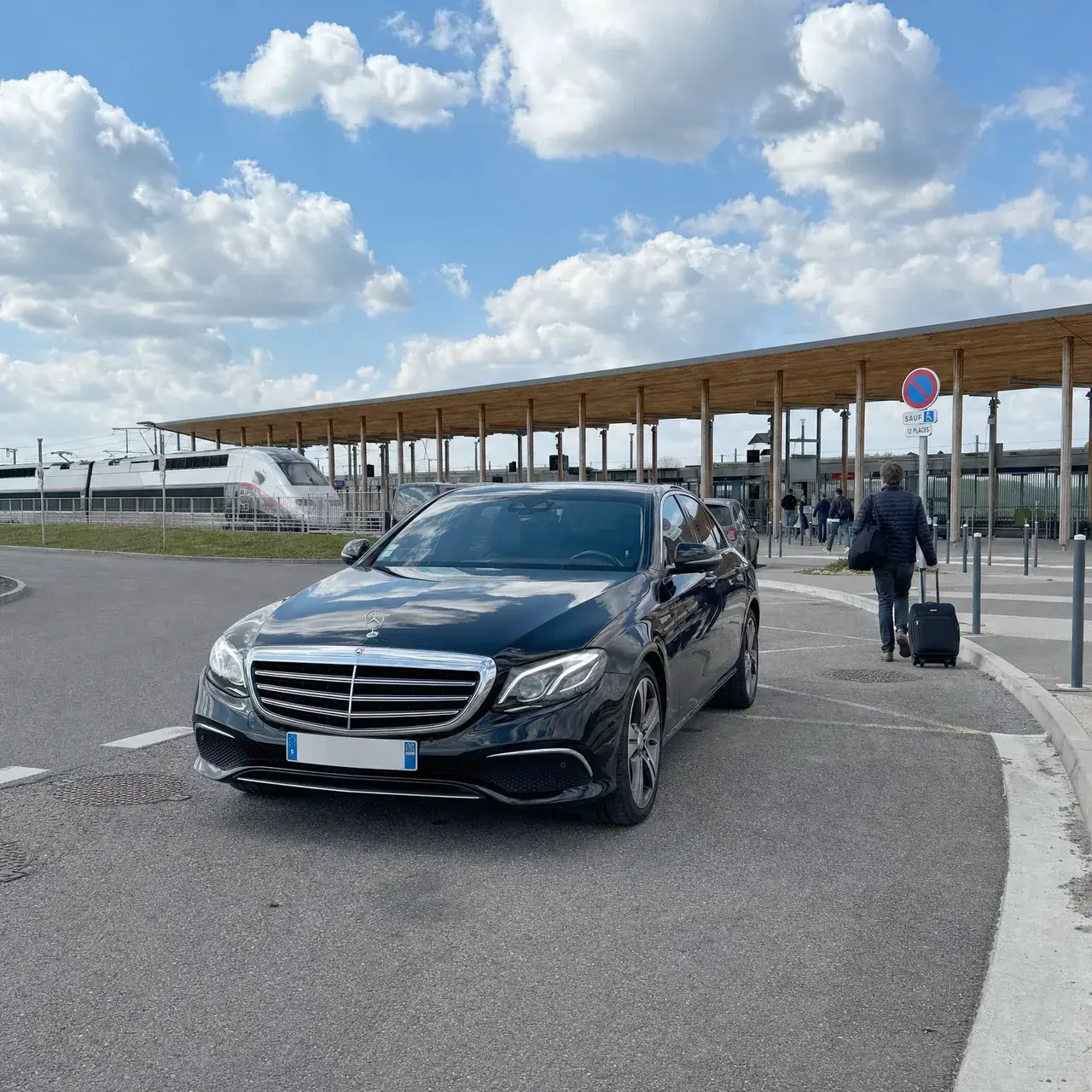 Une Mercedes Classe E noire à la dépose de la gare Lorraine TGV, sous sa longue toiture de bois ; un voyageur de dos part vers les quais avec sa valise