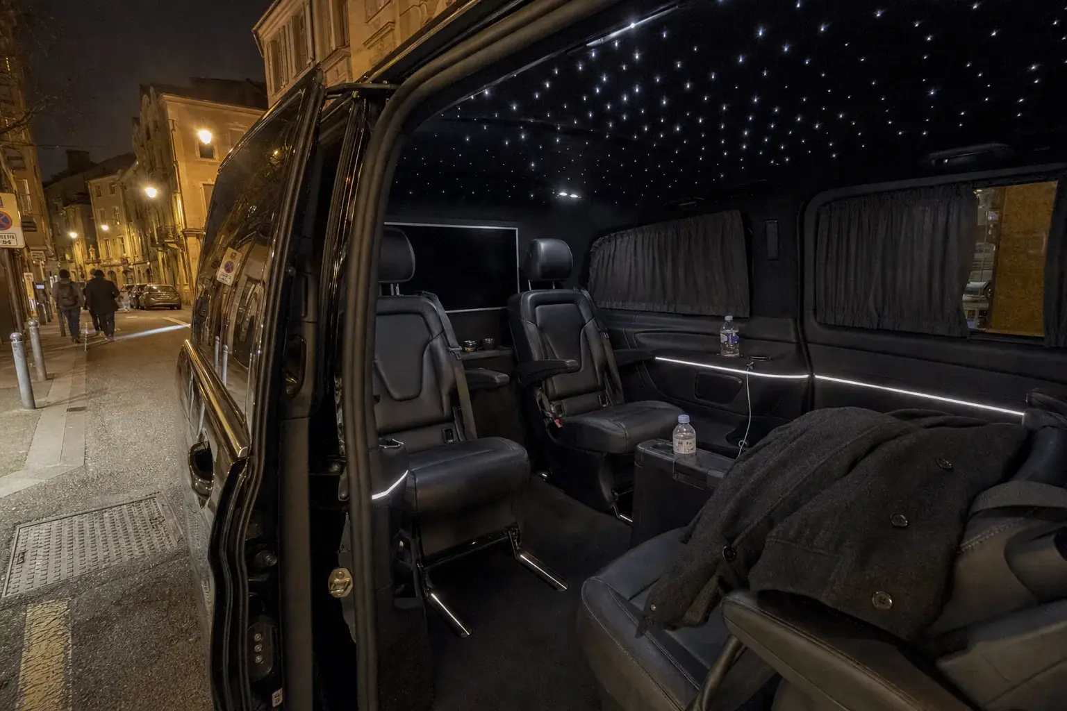 L'arrière d'une Mercedes Classe V noire, portière ouverte le soir dans une rue de Metz : ciel étoilé au plafond, sièges de cuir face à face, rideaux et écran