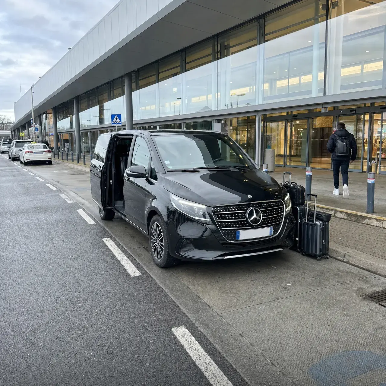 Une Mercedes Classe V noire, portière coulissante ouverte, à la dépose des départs d’un terminal vitré ; des valises sur le trottoir, un voyageur de dos part vers les portes
