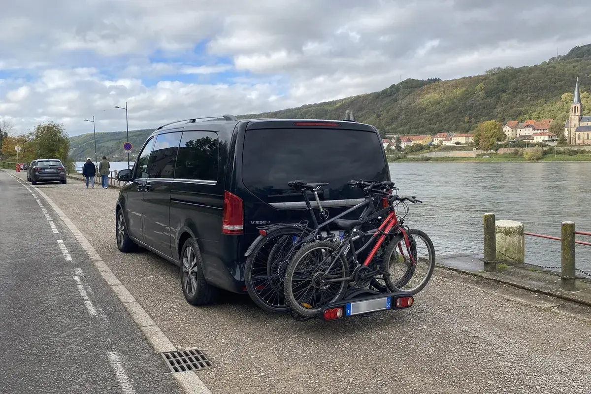 Une Mercedes Classe V noire garée au bord de la Moselle, trois vélos sur son porte-vélos, un village et ses collines en face