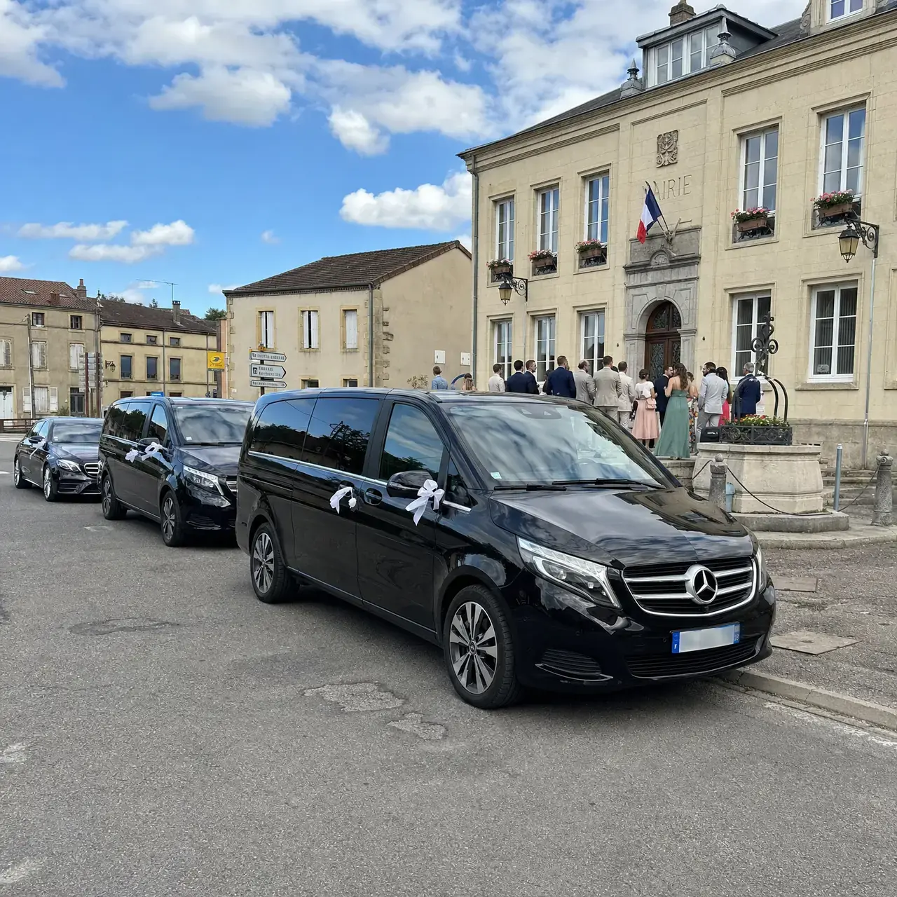 Deux Mercedes Classe V noires et une berline Classe E, rubans blancs aux poignées, garées devant une mairie de village en pierre de Lorraine ; les invités sur le perron