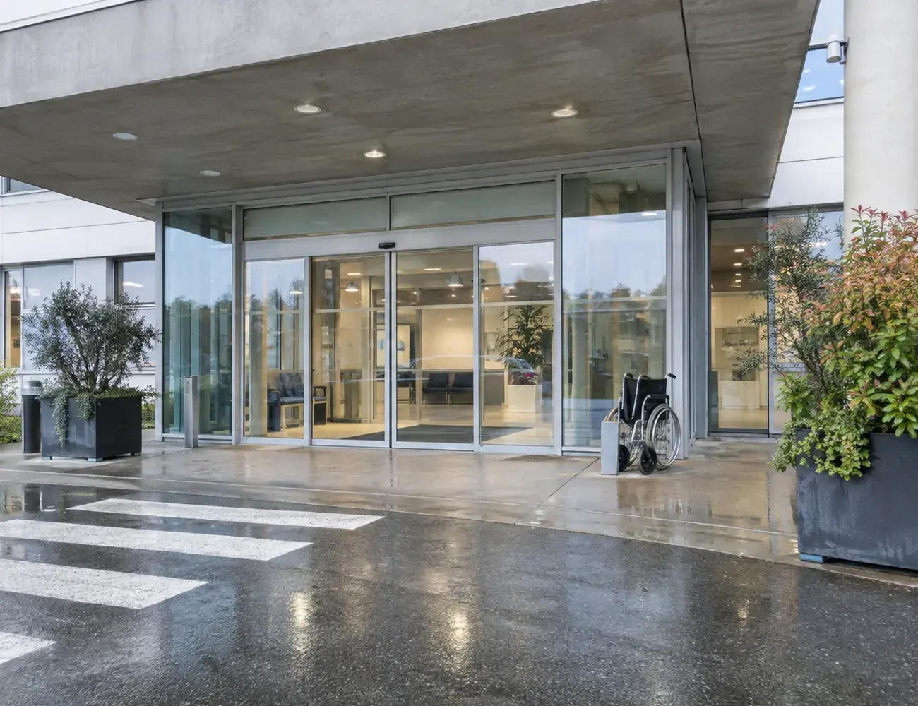 Entrée vitrée d'un hôpital sous une pluie fine, un fauteuil roulant près des portes