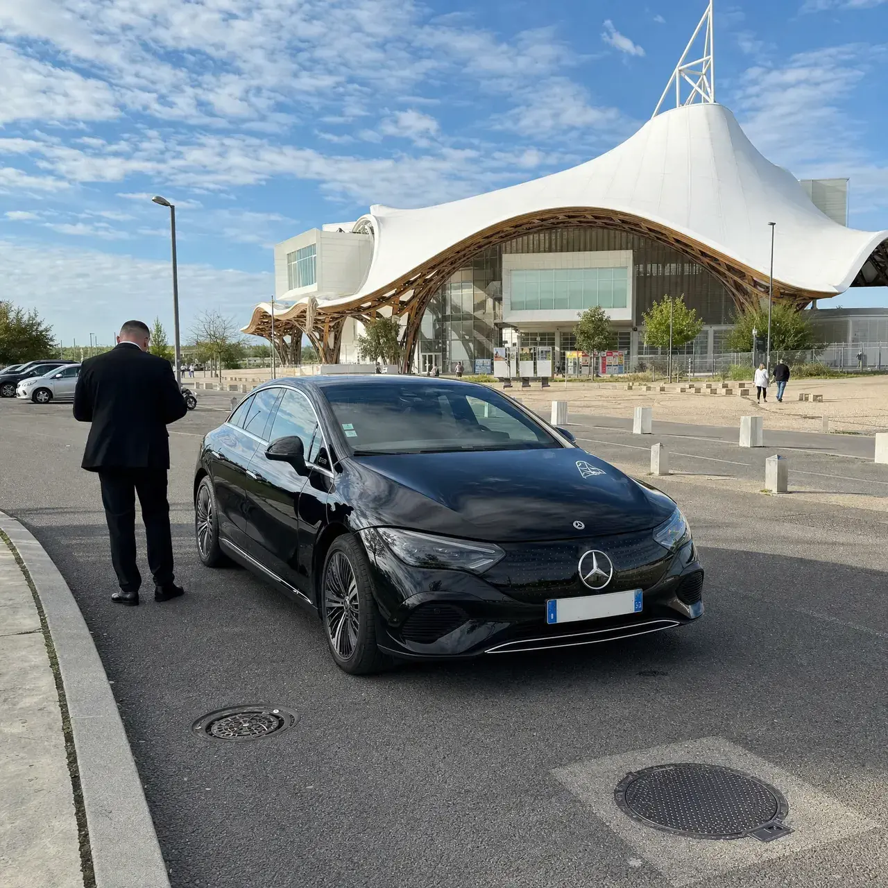 Une Mercedes EQE noire garée devant le Centre Pompidou-Metz ; le chauffeur attend de dos à côté de la portière