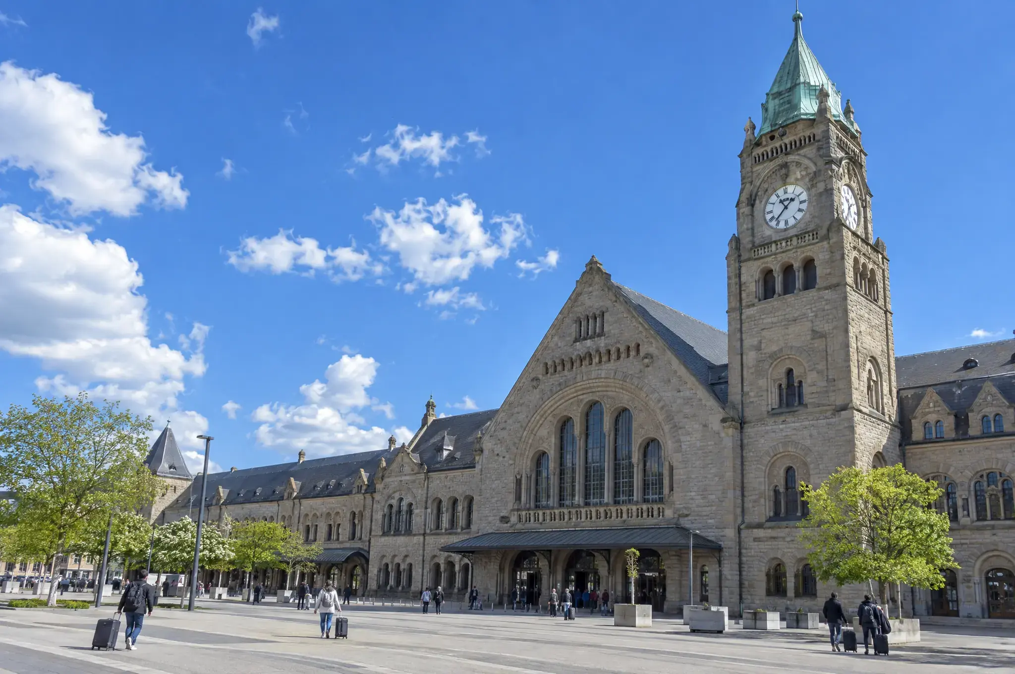 Le campanile à horloge d'une grande gare de pierre, voyageurs et valises sur le parvis, ciel bleu