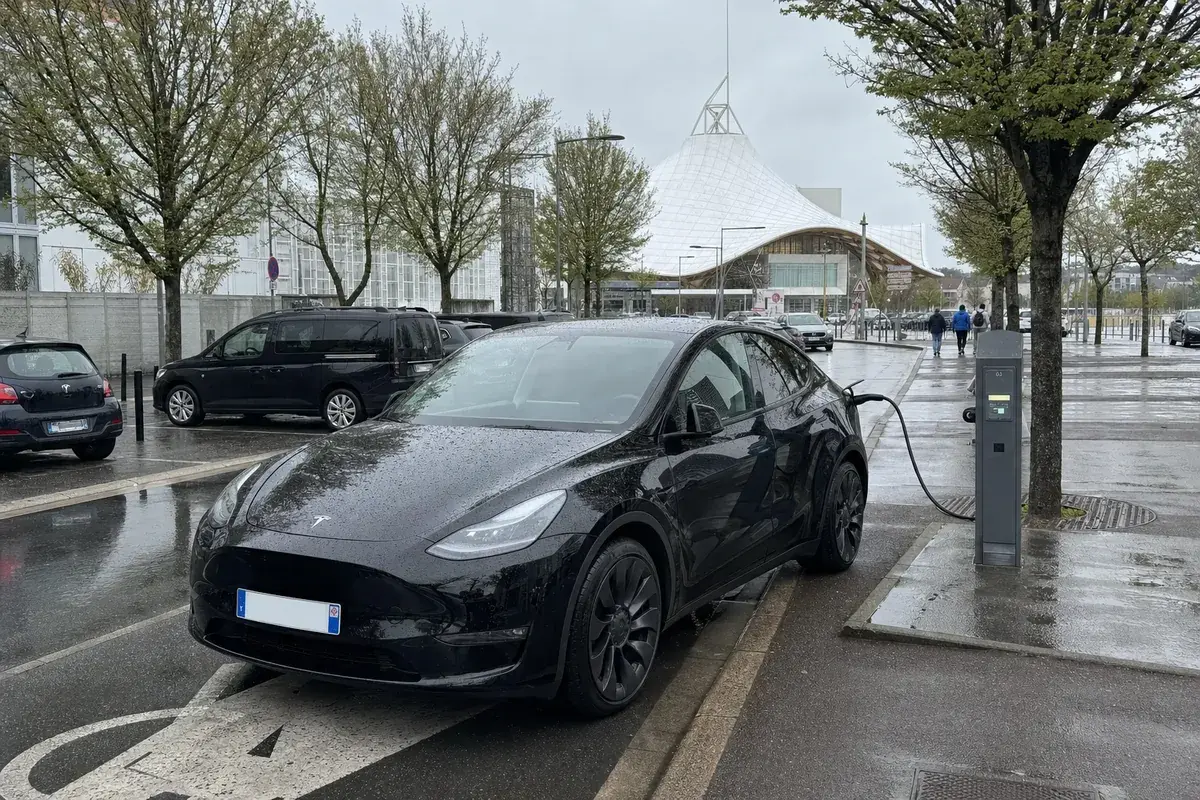 Une Tesla Model Y noire en recharge sur une borne publique, le toit blanc du Centre Pompidou-Metz au fond, chaussée mouillée