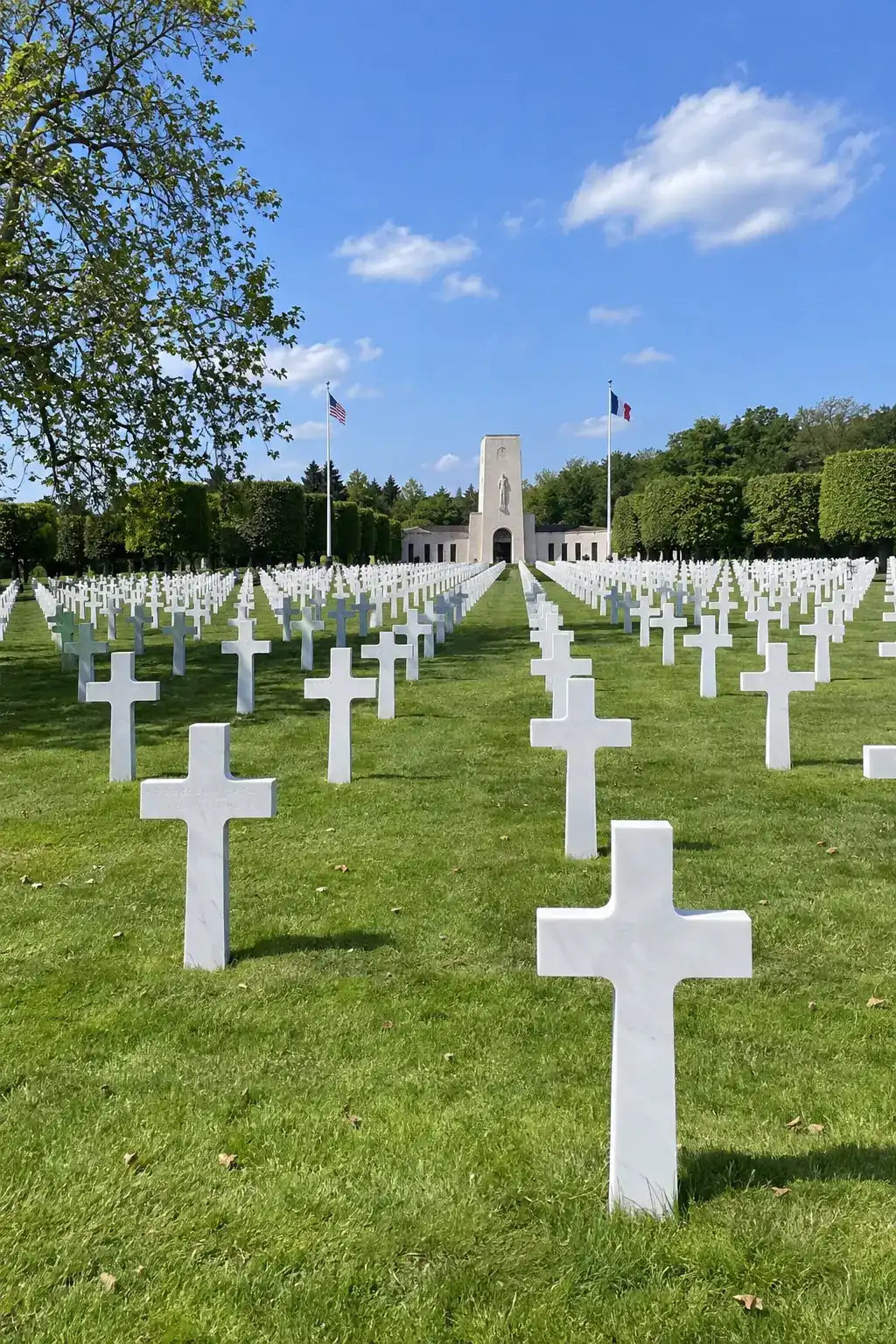 Des rangées régulières de croix de marbre blanc sur une pelouse, une chapelle au loin, journée de printemps