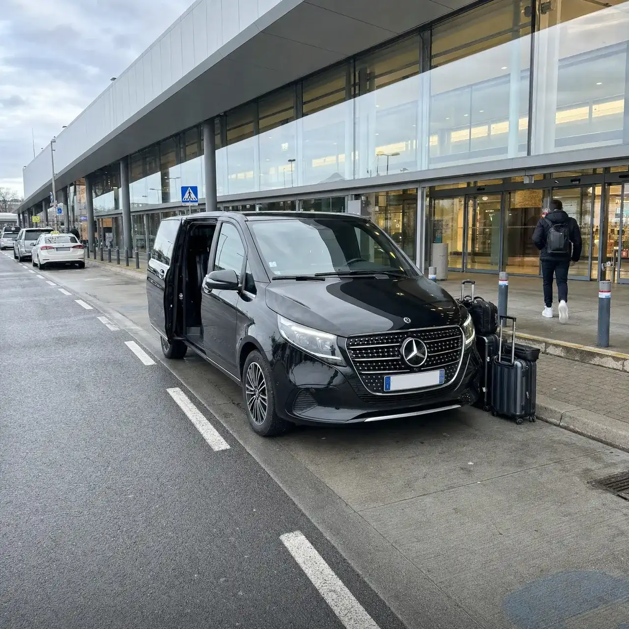 Une Mercedes Classe V noire, portière coulissante ouverte, à la dépose des départs d’un terminal vitré ; des valises sur le trottoir, un voyageur de dos part vers les portes
