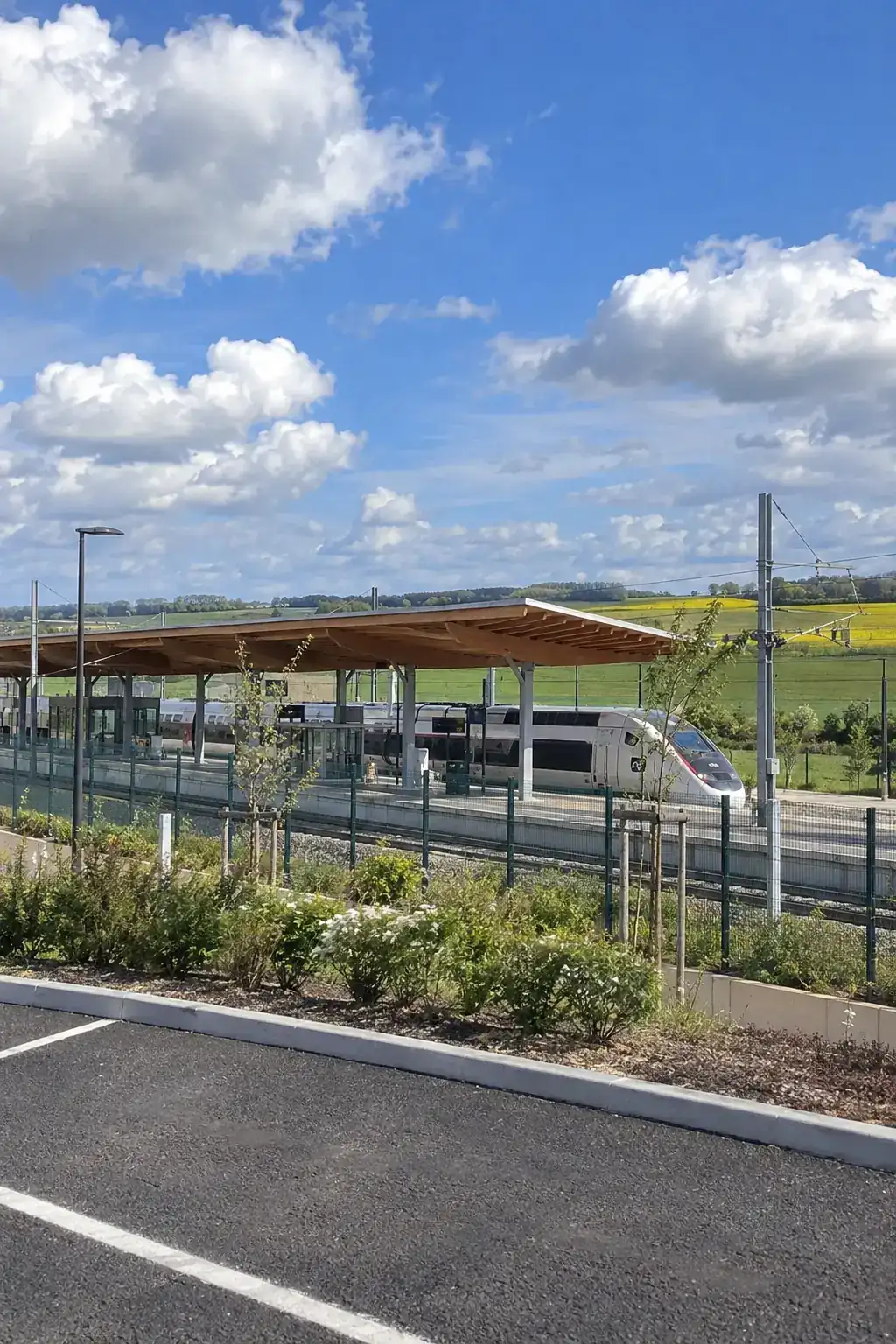Une gare TGV moderne à longue toiture de bois au milieu des champs, un TGV à quai sous un ciel bleu à grands nuages